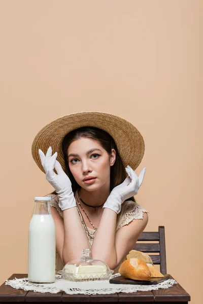 Pretty woman in sun hat and gloves looking at camera near bread and milk on table isolated on beige — Foto stock