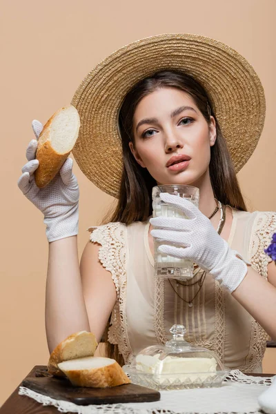 Portrait of stylish woman in sun hat holding milk and bread near butter isolated on beige — Fotografia de Stock