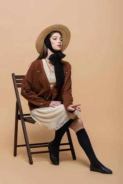 Fashionable model in straw hat posing on chair on beige background — Foto stock