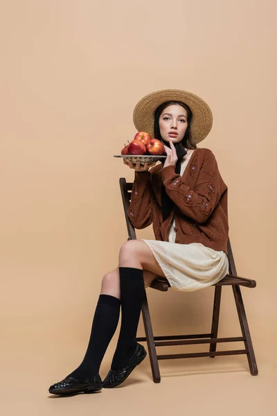 Модна жінка в сонячному капелюсі тримає свіжі яблука, сидячи на стільці на бежевому фоні — стокове фото