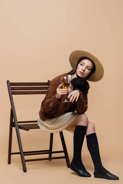 Trendy young woman in straw hat holding wine while sitting on chair on beige background - foto de stock