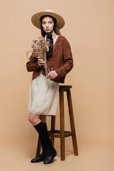 Full length of trendy woman in straw hat holding flowers near chair on beige background — Fotografia de Stock