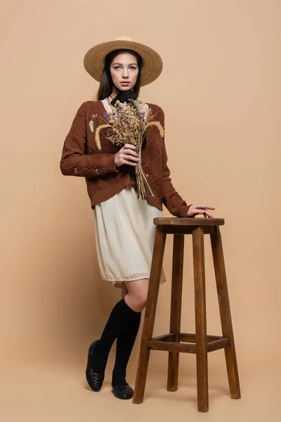 Young woman in straw hat holding plants and looking at camera near chair on beige background - foto de stock