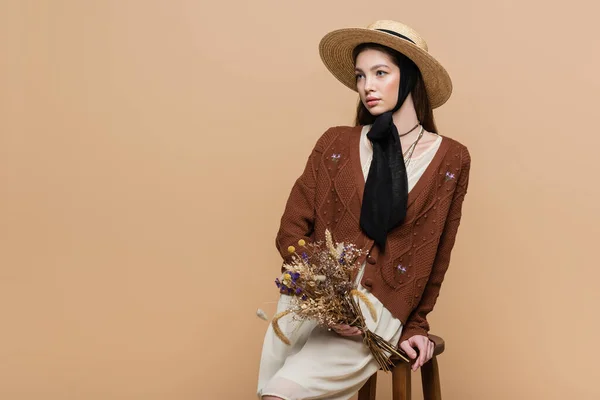 Brunette woman in straw hat holding dry flowers near chair isolated on beige — Stockfoto