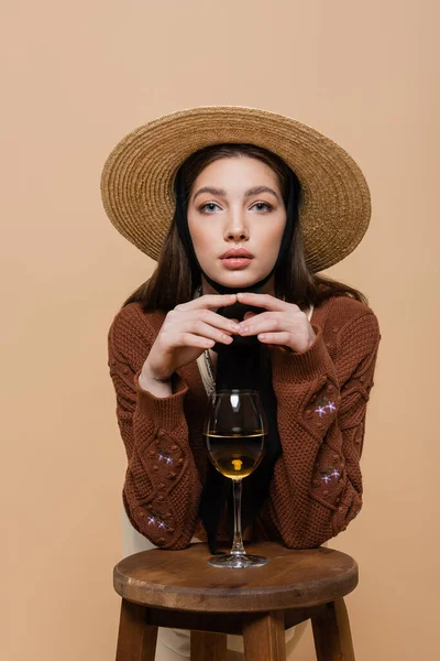 Портрет молодой женщины в шляпе, смотрящей в камеру рядом с вином на стуле, изолированном на бежевом — стоковое фото