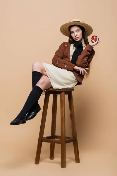Стильна жінка в солом'яному капелюсі тримає яблуко, сидячи на стільці на бежевому фоні — стокове фото