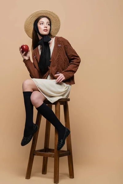 Trendy woman in cardigan and straw hat holding ripe apple while sitting on chair on beige background — Foto stock