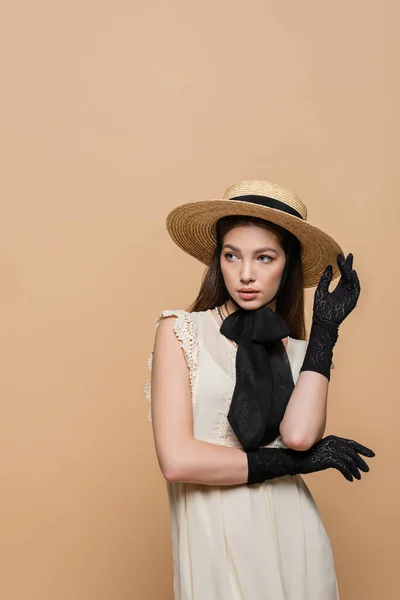 Красивая женщина в соломенной шляпе и черных перчатках позирует изолированно на бежевом — стоковое фото
