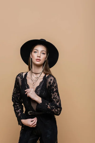 Fashionable woman in black hat and guipure robe looking at camera isolated on beige — Stockfoto