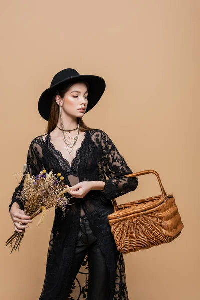Stylish woman in fedora hat holding wicker basket and dry flowers isolated on beige — Foto stock