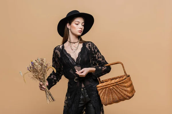 Trendy woman in fedora hat holding wicker basket and plants isolated on beige - foto de stock