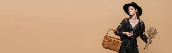 Young woman in black fedora hat holding wicker basket and dry flowers isolated on beige, banner — Foto stock