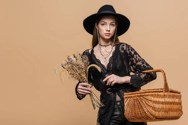 Trendy woman in fedora hat holding dry flowers and wicker basket isolated on beige — Stock Photo