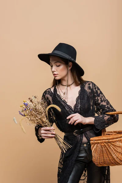 Young woman in guipure robe and fedora hat looking at dry flowers and holding basket isolated on beige - foto de stock