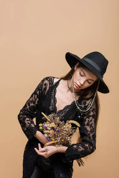 Fashionable woman in fedora hat and guipure robe holding dry flowers isolated on beige — Stockfoto