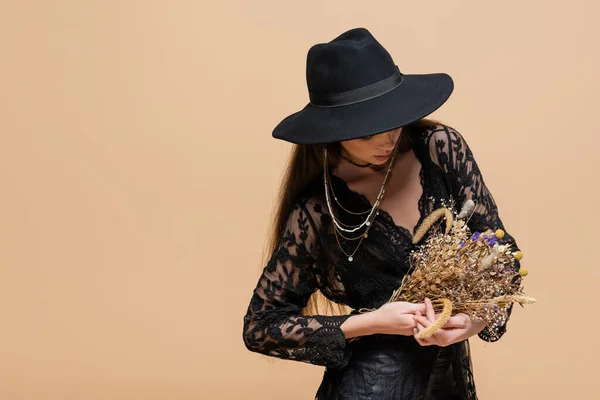 Модна жінка в чорному капелюсі, що тримає рослини, ізольовані на бежевому — стокове фото