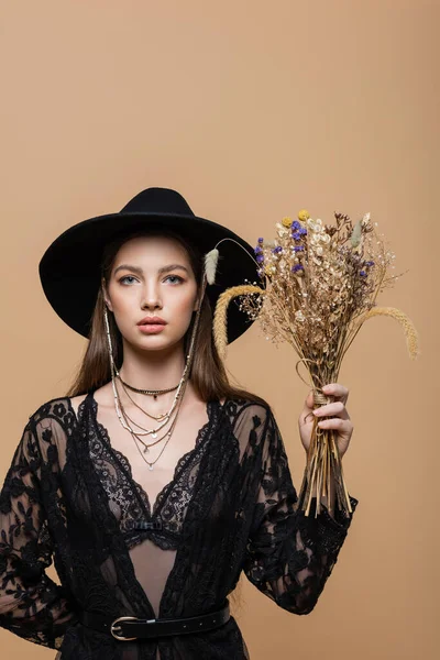 Trendy woman in fedora hat holding plants and looking at camera isolated on beige — Stock Photo