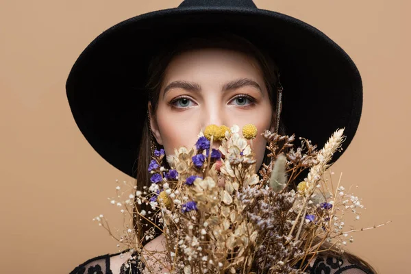 Young woman in fedora hat looking at camera near dry flowers isolated on beige — Stock Photo