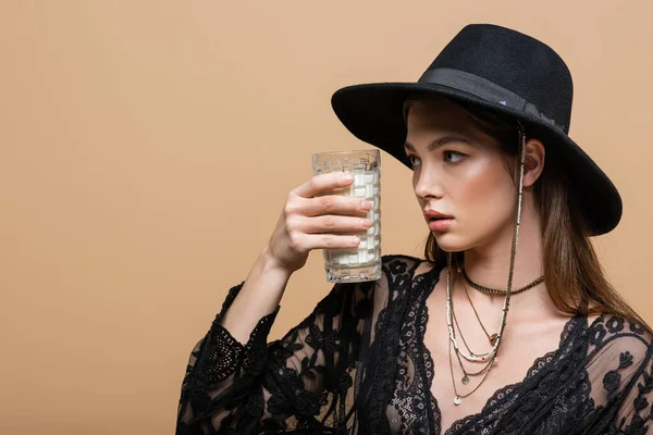 Trendy woman in fedora hat and guipure robe looking at glass of milk isolated on beige — Stock Photo