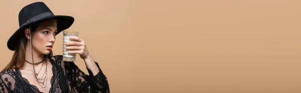 Brunette woman in fedora hat looking at glass of milk isolated on beige, banner — Foto stock