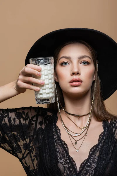 Portrait of fashionable model in fedora hat holding glass of milk isolated on beige — Fotografia de Stock