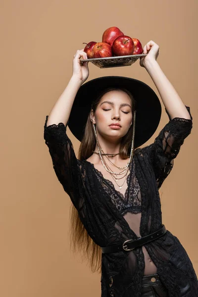 Trendy young woman in black fedora hat closing eyes while holding fresh apples isolated on beige — Photo de stock