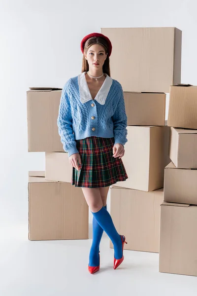 Stylish woman in beret and skirt looking at camera near carton boxes on white background — Stock Photo