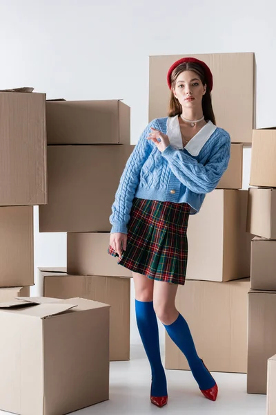 Young woman in plaid skirt and knitted cardigan standing near carton boxes on white background — Stockfoto