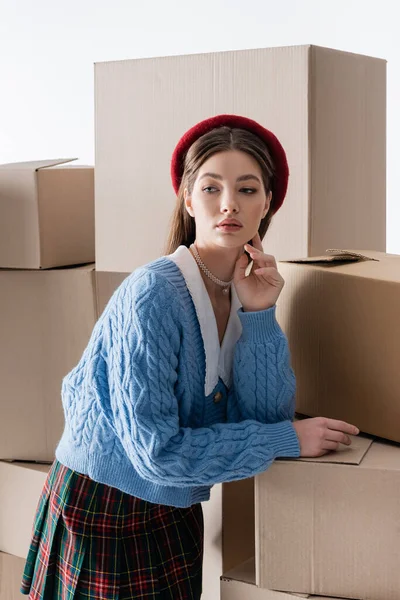 Portrait of stylish woman in beret and knitted cardigan standing near near carton boxes isolated on grey — Fotografia de Stock