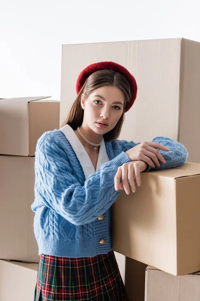 Fashionable woman in beret and cardigan looking at camera near carton boxes isolated on grey - foto de stock
