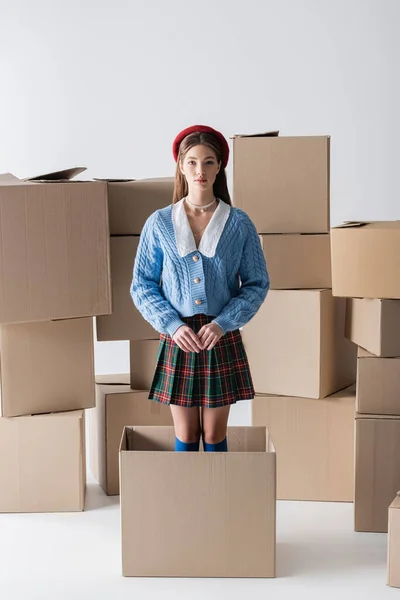 Brunette woman in beret and knitted cardigan standing in package near cardboard boxes on white background — Stockfoto