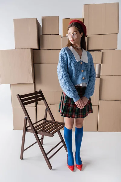 Fashionable model in eyeglasses and beret looking away near chair and cardboard boxes on white background — Stock Photo