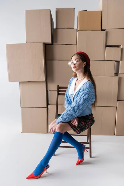 Full length of trendy woman in beret and eyeglasses sitting on chair near cardboard boxes on white background — Foto stock