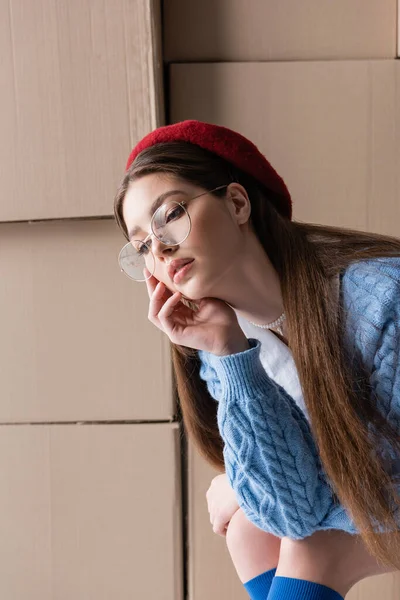 Trendy young woman in eyeglasses and beret posing near cardboard boxes at background — Stock Photo