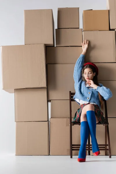 Fashionable woman in eyeglasses and heels sitting on chair near cardboard boxes on white background — Photo de stock