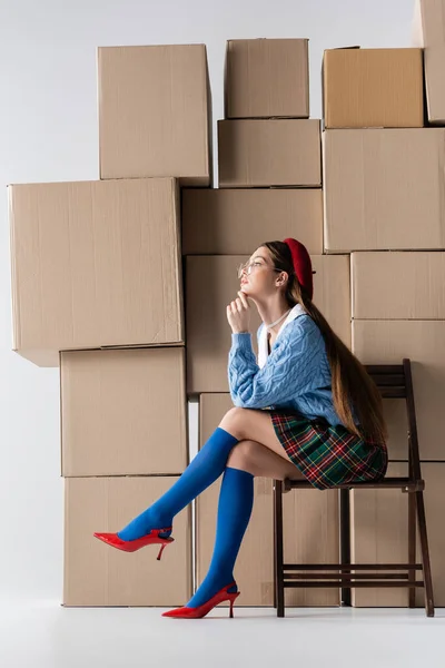 Side view of brunette model in beret and checkered skirt sitting on chair near cardboard boxes on white background — Stockfoto