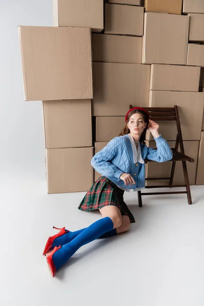 Stylish woman in heels and plaid skirt sitting near chair and cardboard boxes on white background — Foto stock