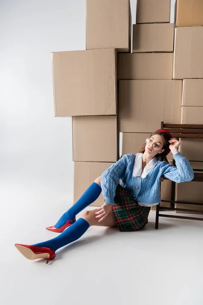 Trendy woman in eyeglasses and beret sitting near cardboard boxes and chair on white background — Photo de stock