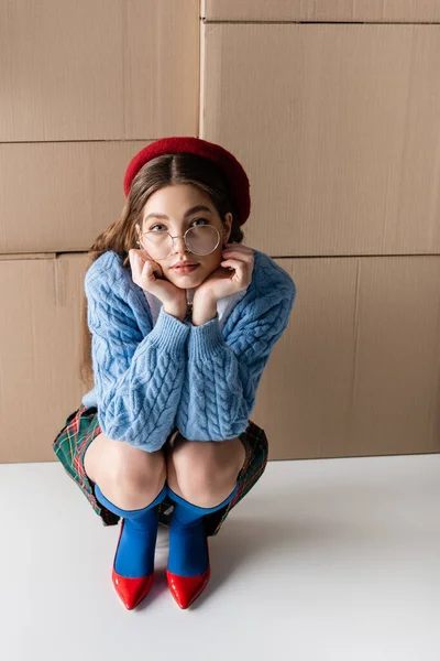 Young model in beret and eyeglasses looking at camera near cardboard boxes on white background — Stockfoto