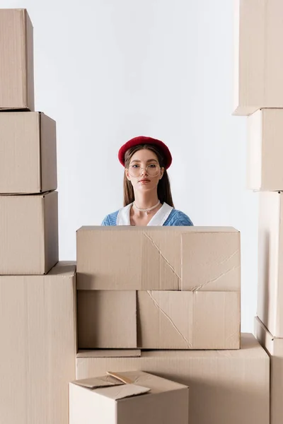 Trendy model in beret and eyeglasses looking at camera behind cardboard boxes isolated on white — Stockfoto