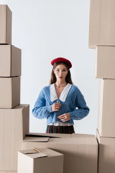 Pretty young woman in eyeglasses and beret touching knitted cardigan near cardboard boxes isolated on white — Stock Photo