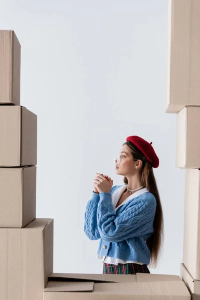 Side view of brunette woman in beret and eyeglasses standing behind cardboard boxes isolated on white — Stockfoto