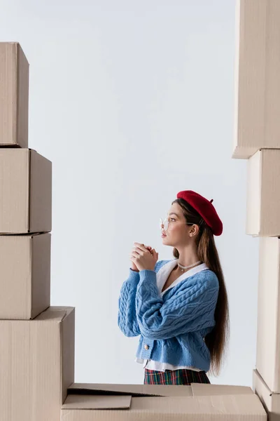 Side view of brunette woman in beret and eyeglasses standing behind cardboard boxes isolated on white — Stock Photo