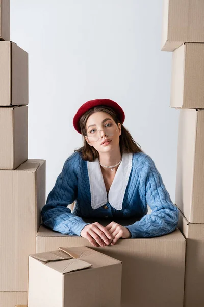 Stylish woman in beret looking at camera between cardboard boxes isolated on white — Foto stock
