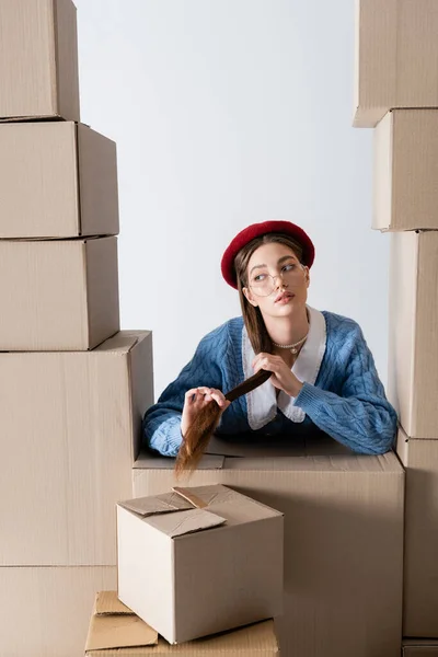 Young model in beret and eyeglasses touching hair between cardboard boxes isolated on white — Stockfoto