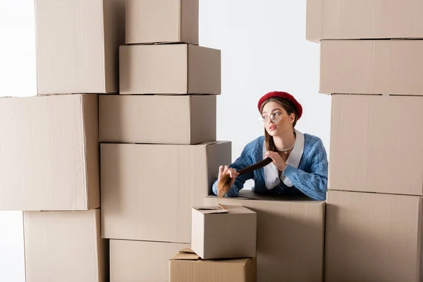 Trendy woman in eyeglasses and beret looking away between cardboard boxes isolated on white — Photo de stock