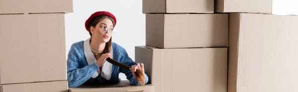 Pretty woman in cardigan and beret touching hair between cardboard boxes isolated on white, banner — Foto stock