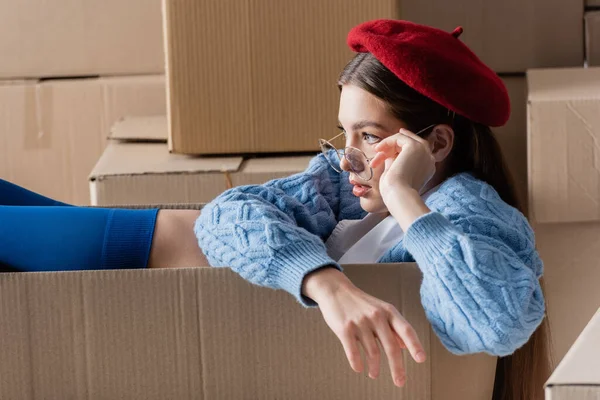 Pretty young model in eyeglasses and beret sitting in cardboard box — Fotografia de Stock
