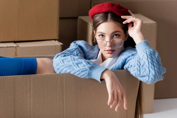 Woman in eyeglasses and cardigan sitting in package and looking at camera near cardboard boxes on white background — Photo de stock