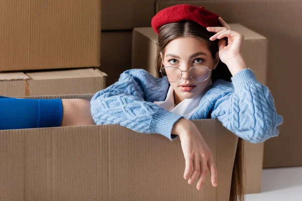 Woman in eyeglasses and cardigan sitting in package and looking at camera near cardboard boxes on white background — Stockfoto